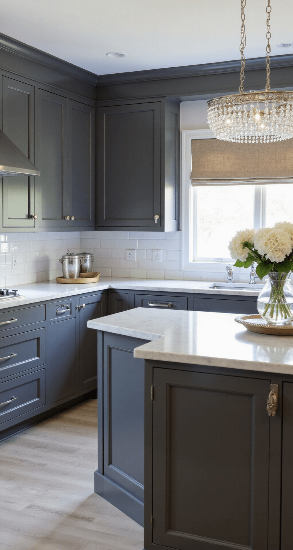 Traditional gray kitchen with deep graphite shaker cabinets, Carrara marble countertops, and polished nickel hardware, illuminated by early morning light. Features crown molding, raised panel details, and an elegant chandelier over the island, styled with white hydrangeas and silver serving pieces.