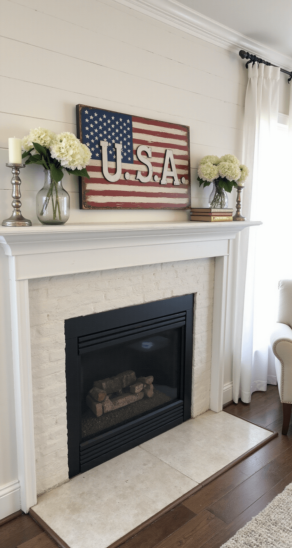A vintage Americana living room mantel styled with a hand-painted distressed wooden USA sign, vintage books, mercury glass candlesticks, and white hydrangeas, set against a classic white shiplap wall, illuminated by soft afternoon light filtering through sheer curtains.