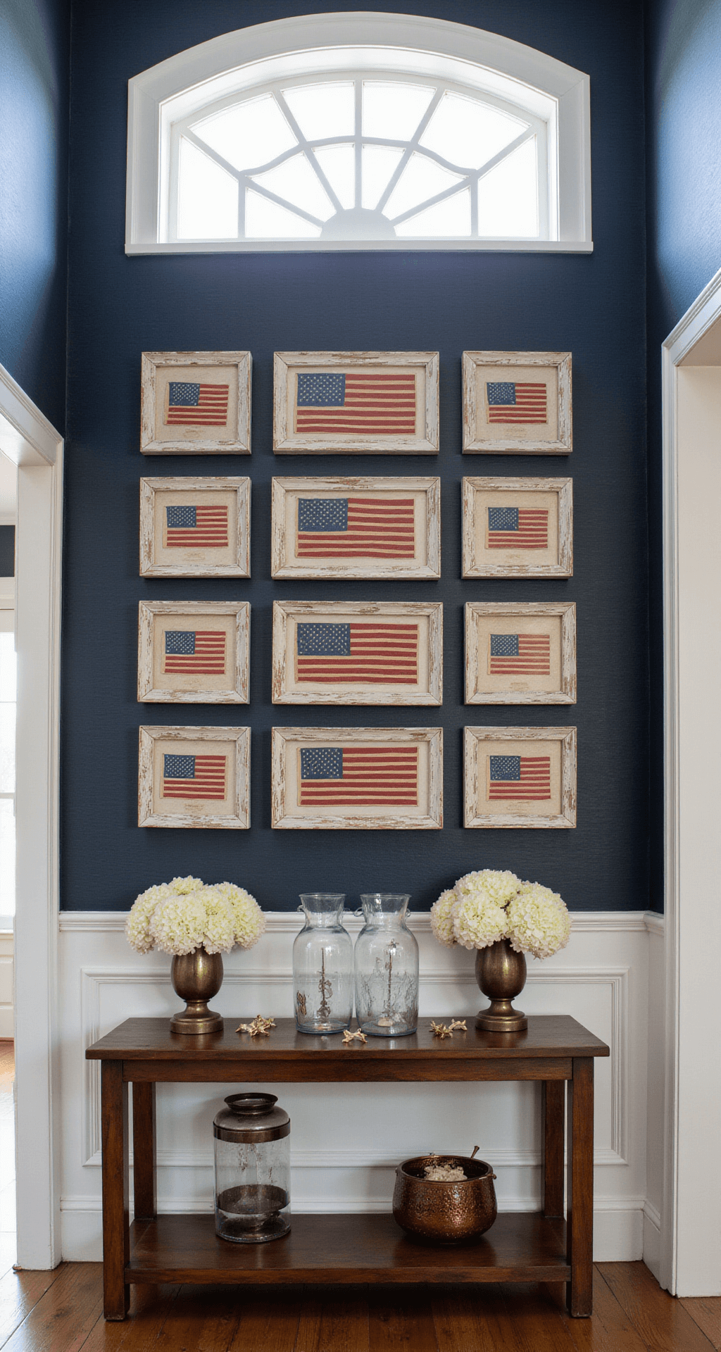 A sophisticated foyer featuring a gallery of historical American flags framed in distressed wood, a console table with mercury glass hurricanes and white hydrangeas, against navy grasscloth wallpaper, illuminated by soft morning light through a transom window.