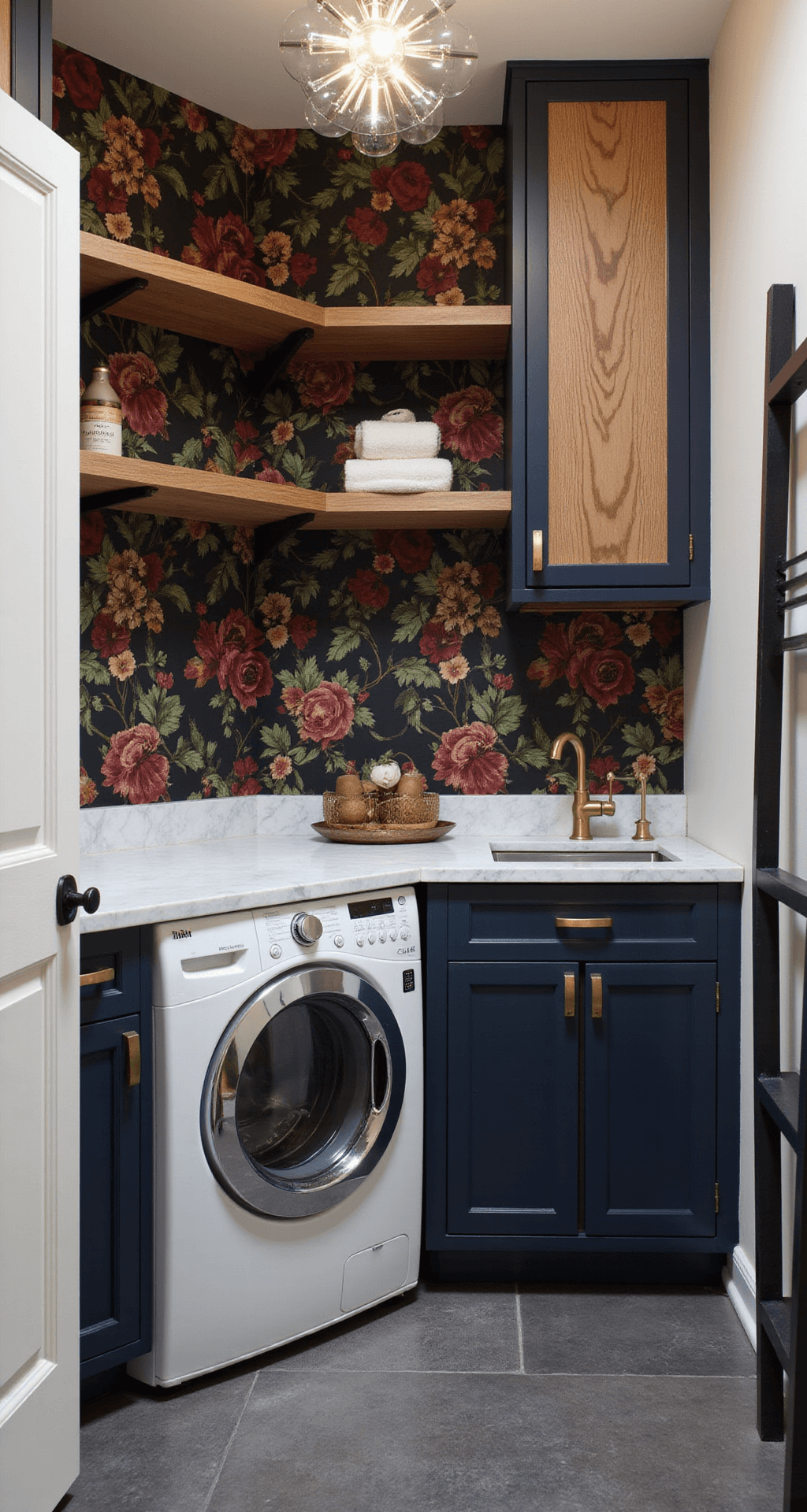 A stylish laundry room with deep navy cabinets and brass hardware surrounding a white washer and dryer, adorned with floral wallpaper in burgundy, gold, and green on a black background. A marble countertop and two-tone shelving units enhance the space, complemented by a black ladder rack and charcoal tiled flooring. Modern ambient lighting from a sputnik chandelier creates intriguing shadows in this vibrant, compact area.