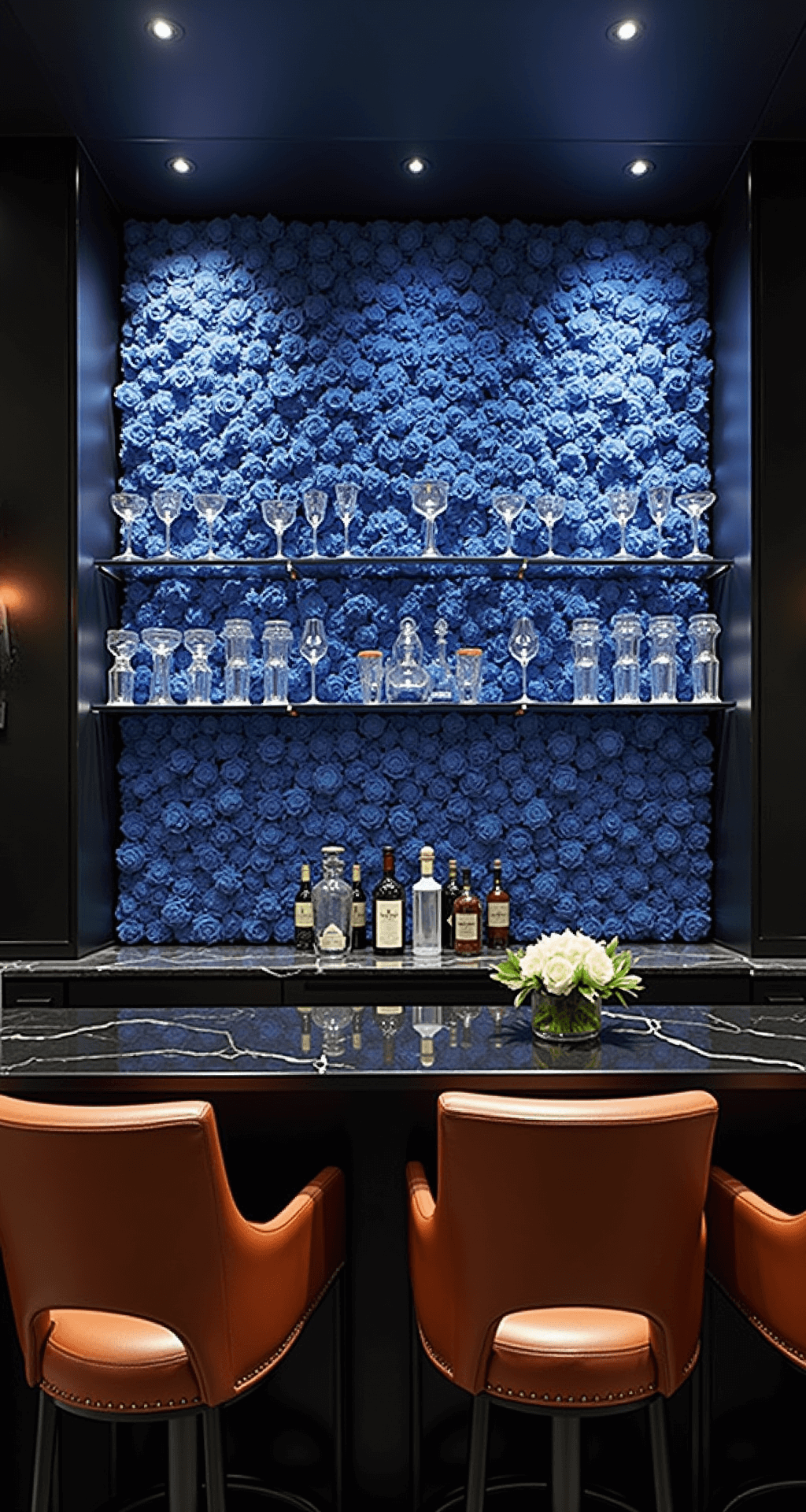 A sophisticated home bar area featuring a striking backsplash of pipe cleaner flowers in deep blues and indigos, complemented by a sleek black marble countertop and rich cognac leather barstools, all illuminated by low ambient lighting with spotlights on the floral installation.