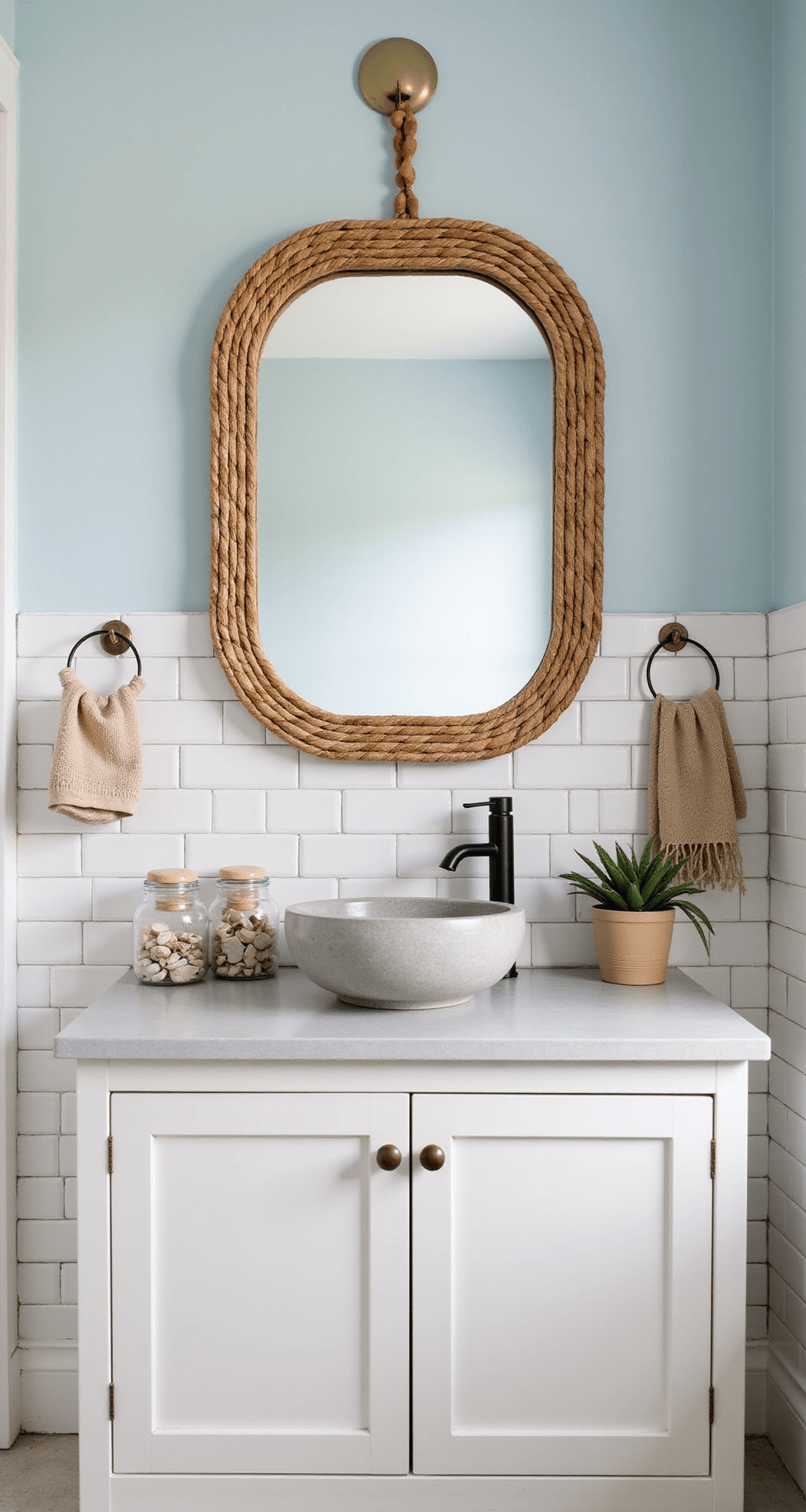 A coastal boho bathroom vanity styled in morning light, featuring a rattan-framed mirror over a concrete vessel sink, white subway tile backdrop, light blue walls, glass canisters with shells, wooden beads, and hand-woven turkish towels in sandy tones, accented by a small potted succulent.