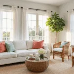 Photorealistic interior of a sunlit coastal living room featuring a white linen sofa with coral and seafoam green pillows, vintage rattan armchairs, a natural seagrass rug, and jute rope wall art, all bathed in golden hour light through floor-to-ceiling bay windows.