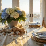 A sunlit coastal dining room at golden hour featuring a weathered teak table with layered place settings, a natural linen runner, white ceramic plates on rattan chargers, a centerpiece of fresh hydrangeas and driftwood, and large windows with sheer curtains.
