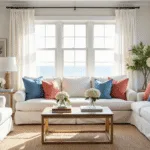 Hyperrealistic image of a sun-drenched coastal living room featuring a white linen sofa with coral and azure pillows, large bay windows, a jute rug, brass accents, and fresh white hydrangeas, showcasing a perfect blend of cool ocean blues and warm sandy neutrals.