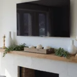 Cinematic interior of a sunlit coastal living room featuring a limestone fireplace with a white oak mantle, Samsung Frame TV, and stylish decor including ceramic vases, eucalyptus garland, and brass candlesticks, all in a palette of whites, blues, and greens.