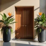 A modern entryway with a 12-foot ceiling, featuring two matte black cylindrical planters with 6-foot Bird of Paradise plants, surrounded by Coleus and English Ivy, dramatic afternoon lighting, and polished concrete flooring.