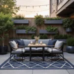A cozy urban patio at golden hour featuring a geometric navy and cream rug, a sleek black metal bistro set with grey cushions, and a weathered teak ottoman, surrounded by modern wall planters with cascading greenery and warm string lights overhead.