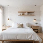 Photorealistic image of a bright and airy 10x12ft bedroom featuring a queen bed with white linens, a textured throw, a white-painted brick accent wall, minimalist oak shelves, and a brass wall sconce, bathed in morning sunlight through sheer curtains, with pale oak flooring and a jute rug, capturing a serene and hygge atmosphere.