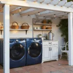 A modern outdoor laundry room in a lush garden, featuring a navy blue washer and dryer under a wooden pergola, a vintage folding station, and galvanized shelves with laundry supplies, illuminated by warm sunlight and surrounded by climbing jasmine and ferns.