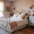 A dreamy vintage bedroom with a distressed white iron bed adorned in soft linen bedding and embroidered pillows, bathed in golden hour light. A weathered French provincial dresser and ornate mirror complement the blush pink walls, while floral rugs and brass sconces enhance the romantic atmosphere.