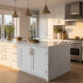 A luxury contemporary kitchen featuring a white Shaker-style island with a Carrara marble waterfall countertop, illuminated by golden hour light through floor-to-ceiling windows, with warm shadows on wide-plank white oak flooring, brass accents, and artisanal decor.