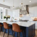 Photorealistic contemporary kitchen featuring a navy blue island with a white marble waterfall countertop, illuminated by natural sunlight, glass pendant lights, white shaker cabinets, oak herringbone flooring, brass hardware, cognac leather barstools, and white hydrangeas.