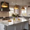 A luxurious kitchen featuring a 12-foot marble island with dramatic grey veining, white shaker cabinets, deep charcoal granite countertops, and oversized brass pendant lights, all illuminated by soft natural light from floor-to-ceiling windows, adorned with fresh hydrangeas and copper cookware.