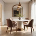 A sophisticated small dining room with a round white marble table, dusty rose velvet chairs, cream walls, and herringbone flooring, illuminated by golden hour light through large windows. An oversized antiqued mirror and a minimalist brass pendant lamp add elegance, while sheer curtains filter sunlight.