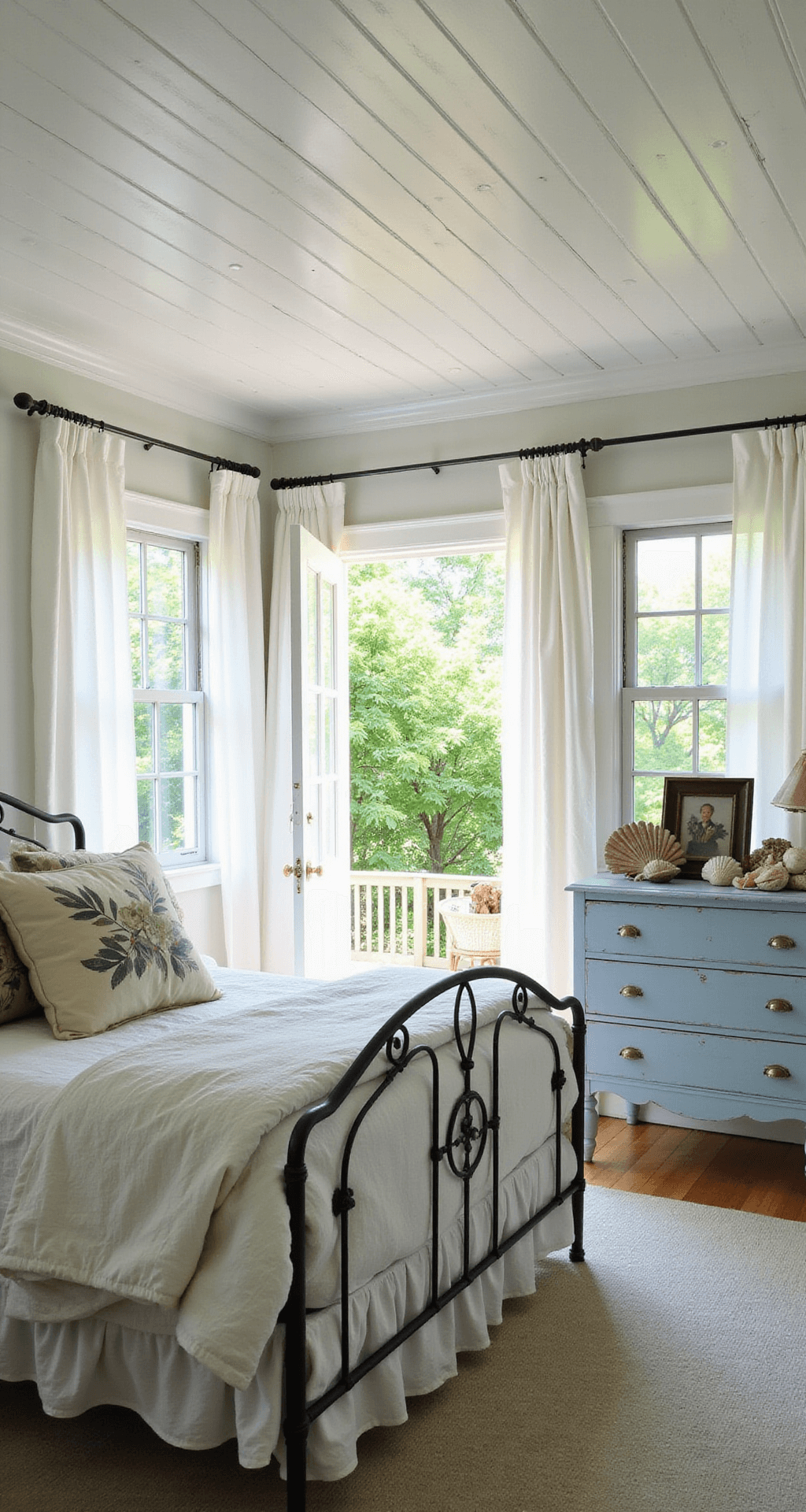 A cozy summer cottage bedroom features a wrought iron bed with vintage linens and botanical pillows, a pale blue dresser adorned with seashells and photographs, and billowing white curtains, all bathed in soft morning light. The doorway provides a wide-angle view of the serene interior contrasting with the lush garden outside.