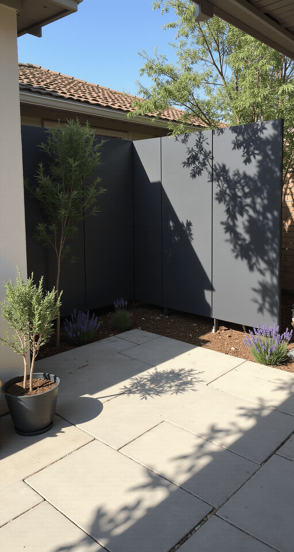 Modern courtyard with zigzagged matte charcoal PVC privacy screens featuring geometric cutouts, casting artistic shadows on light concrete pavers, surrounded by potted olive trees and lavender, captured from a high overhead angle in bright midday light.
