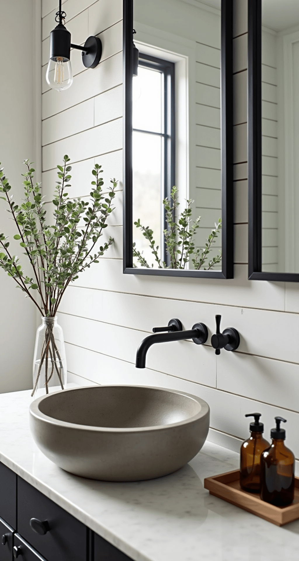 A modern farmhouse bathroom vignette featuring white shiplap walls, a black steel-framed mirror, a concrete vessel sink, eucalyptus branches in a clear glass vase, and vintage amber glass apothecary bottles on a marble tray, illuminated by early morning light.