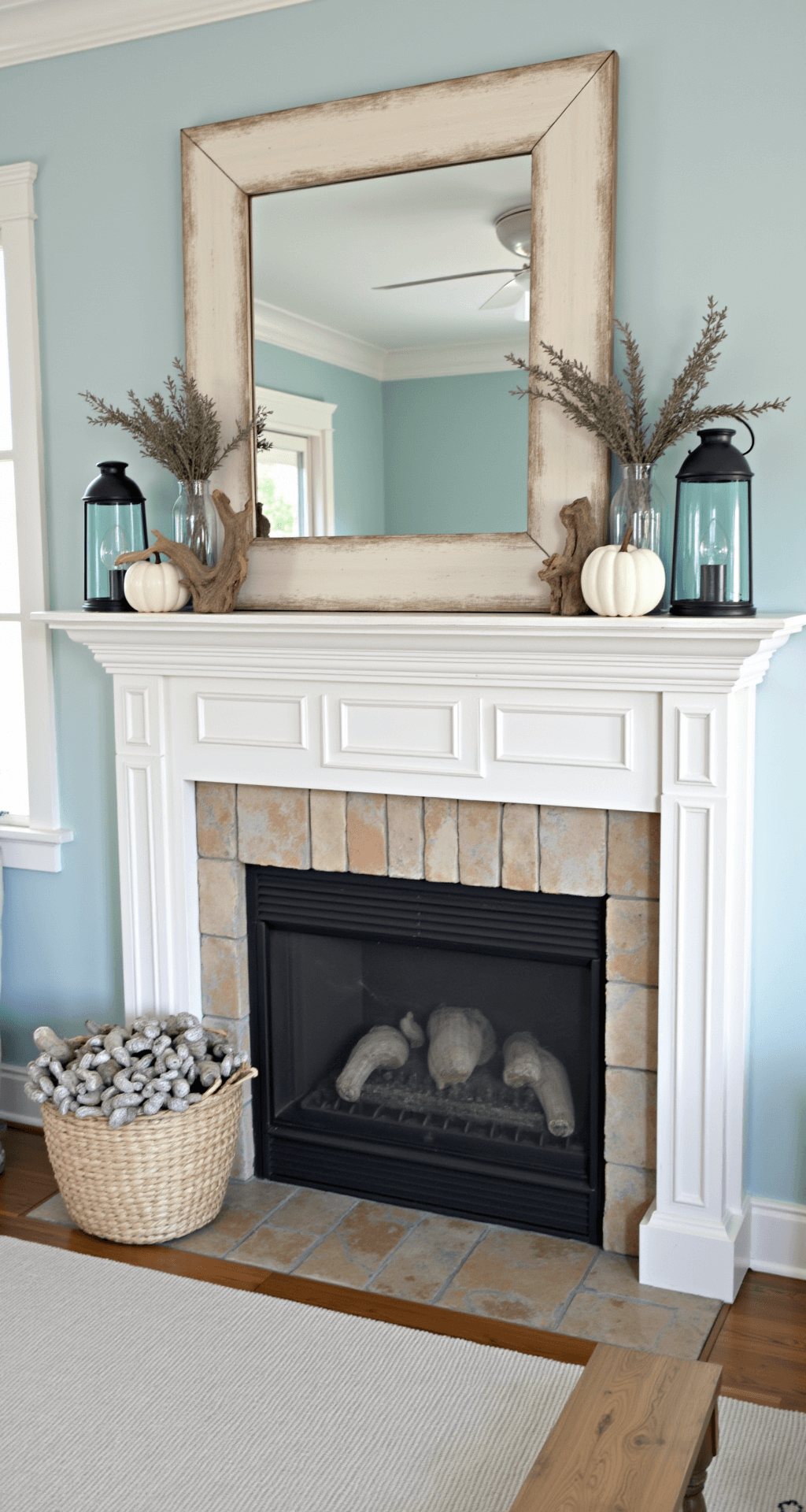 A bright coastal living room with pale blue walls features a whitewashed wood mantel decorated with white ceramic pumpkins, driftwood, dried sea lavender, and blue glass hurricane lanterns. Natural light brightens the space, casting reflections from nearby water and highlighting the layered depth of the decor.