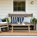 A cozy front porch with a weathered rocking chair and navy striped cushions, rustic bench with a cream cable-knit blanket, galvanized herb planters, Edison bulb string lights, and golden hour lighting, creating a warm and inviting atmosphere.