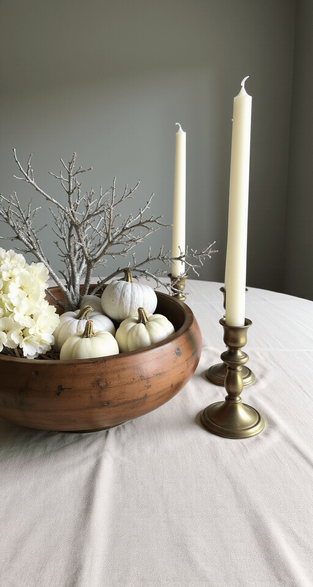 Elegant Thanksgiving Table Centerpieces That'll Make Your Guests Forget About the Turkey A rustic wooden dough bowl filled with white pumpkins, silver-painted branches, preserved hydrangeas, and ivory candles in brass holders, set on a natural linen tablecloth against muted gray-green walls, enhanced by gentle side lighting.