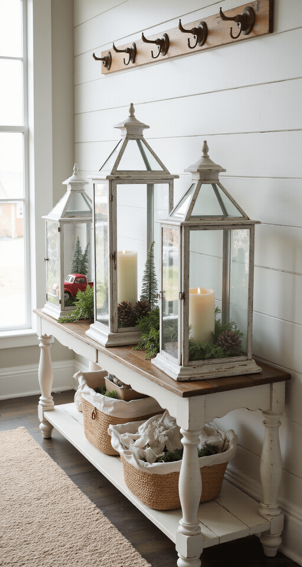 Christmas Lantern Decorating Ideas That Actually Work Elegant farmhouse entryway with a distressed white console table adorned with three glass lanterns, each showcasing unique winter scenes, illuminated by soft natural light. A textured wool runner lies beneath the table, complemented by aged brass hooks on a shiplap wall, evoking warmth and nostalgic charm.