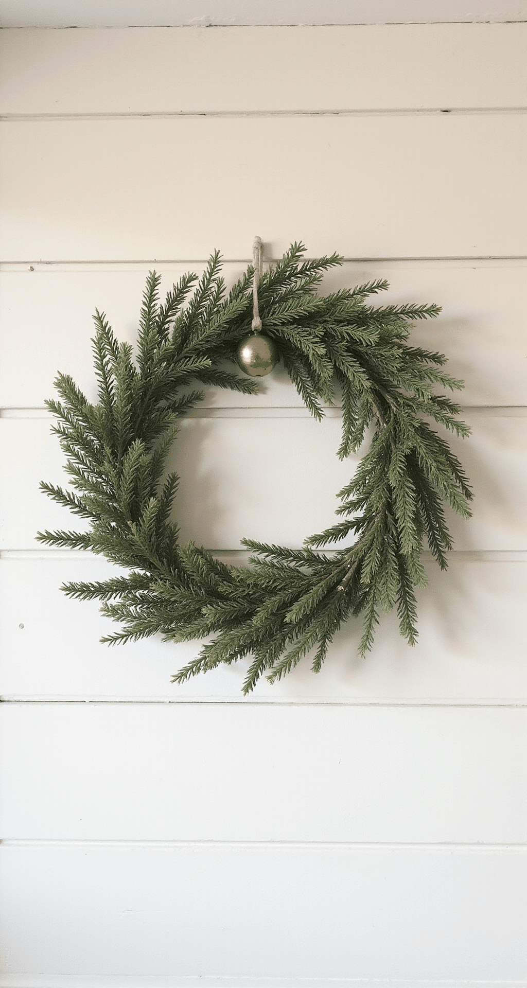 DIY Holiday Wreaths That'll Make Your Front Door the Talk of the Neighborhood (In Under 15 Minutes!) A minimalist farmhouse holiday wreath scene featuring a brass bell-accented natural greenery wreath asymmetric on a soft white shiplap wall, with warm neutral tones, gentle shadows, and soft winter morning light highlighting the textured cedar and hemlock branches in a muted color palette.