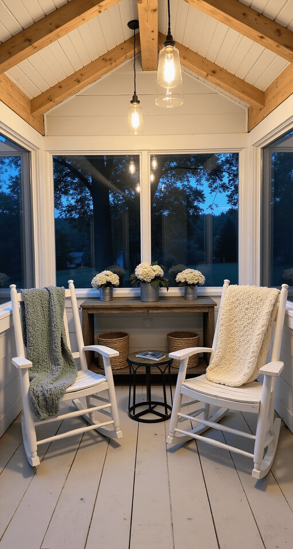 Screened-In Back Porch Designs That'll Make You Actually Want to Go Outside A cozy screened porch with a white shiplap ceiling and exposed pine beams, featuring two white rocking chairs with cream and sage throw blankets, a galvanized metal side table, and a distressed wooden console table with vintage planters of white hydrangeas. Warm ambient light from pendant lamps contrasts with the cool twilight outside, all set on white-washed plank flooring.