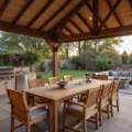 Cinematic outdoor pavilion at golden hour featuring a dramatic gable roof, weathered teak furniture, climbing purple clematis vines, and a cozy fire pit, set against a contemporary home backdrop with lush landscaping.