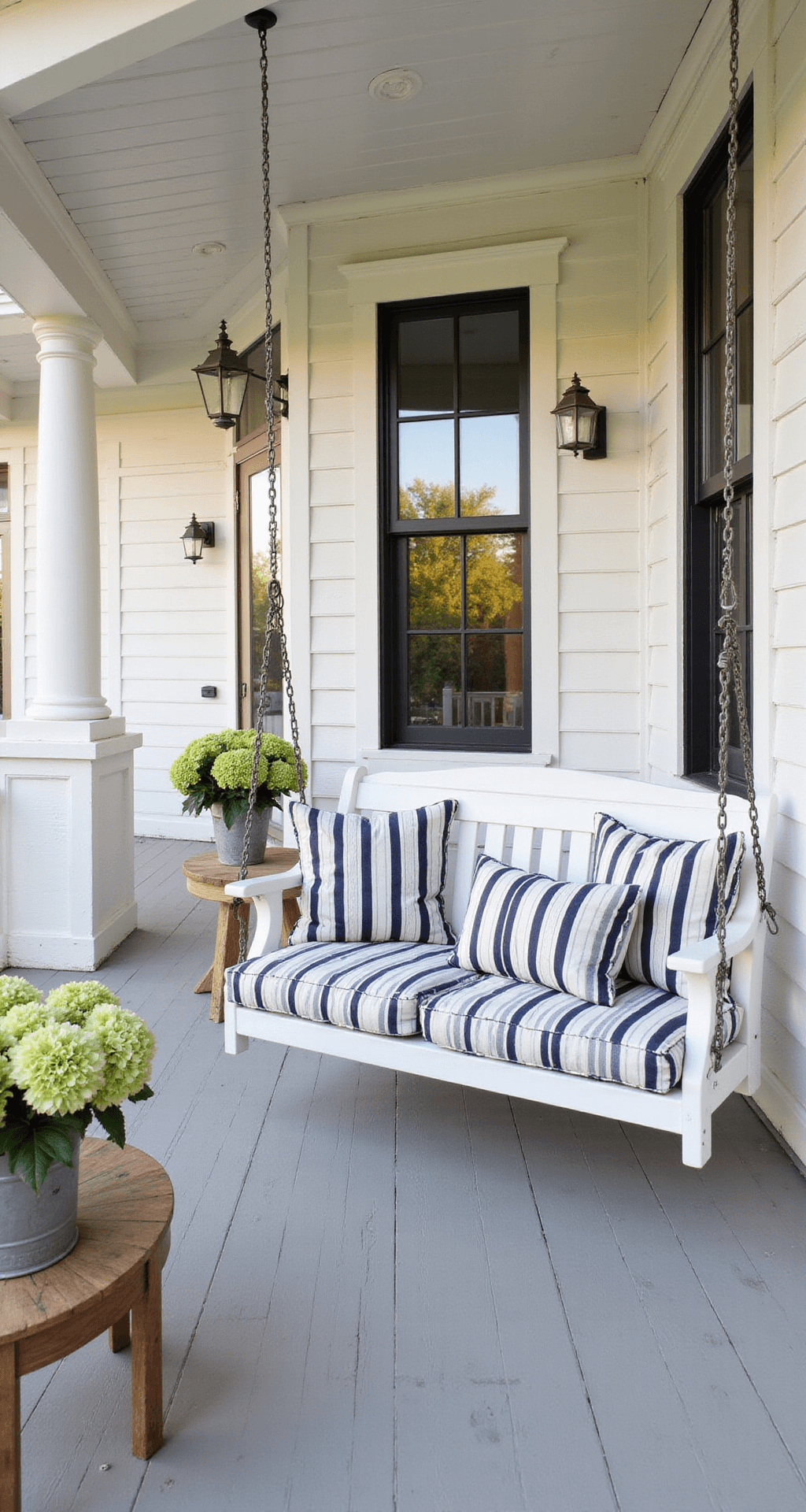 The Front Porch Chair That Made My Neighbors Stop and Stare (And How You Can Find Yours) A modern farmhouse porch with a white wooden swing, navy and white striped cushions, gray painted flooring, and a teak side table with hydrangeas, all bathed in warm late afternoon sunlight.