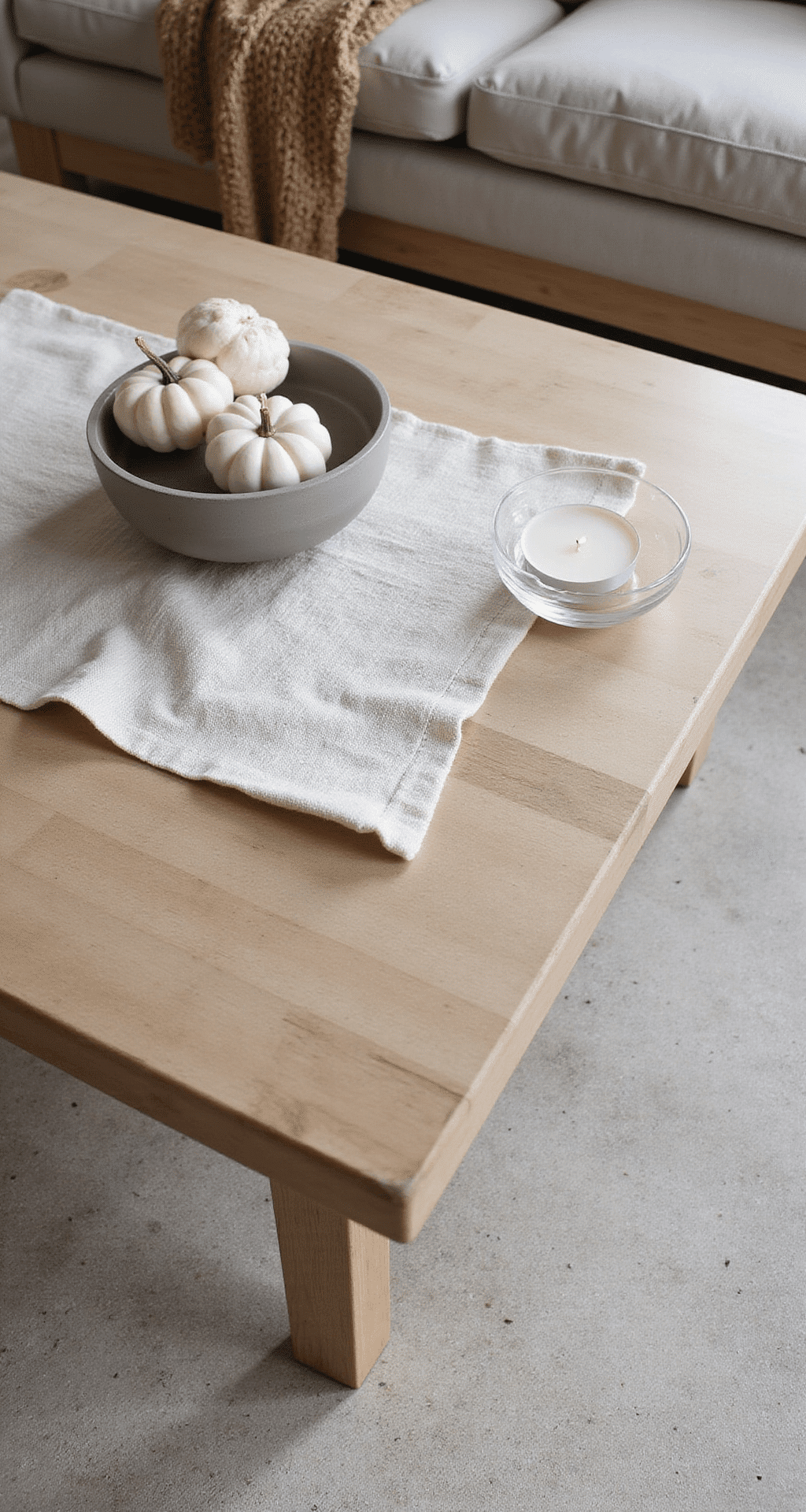 Minimalist Fall Decor: How I Finally Cracked the Code to Cozy Without the Clutter A minimalist coffee table vignette showcasing a bleached oak table with a natural linen runner, featuring a warm gray concrete bowl, three white pumpkins, and a glass tealight holder, illuminated by soft natural light, with a chunky knit camel throw in the background.