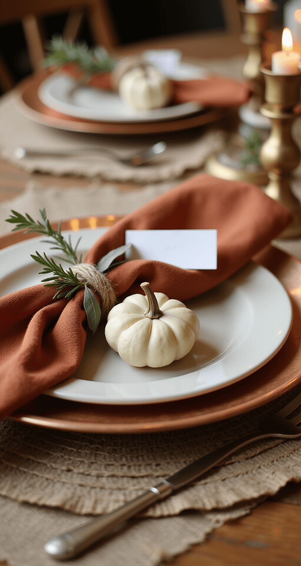 Fall Dining Room Table Decor: Transform Your Space in Under 2 Hours A close-up of a fall tablescape featuring a matte cream ceramic dinner plate on a hammered copper charger, with a terracotta linen napkin tied with jute twine, a sprig of rosemary, and a sage leaf. A small white pumpkin serves as a place card holder, while warm candlelight creates a cozy, intimate atmosphere.